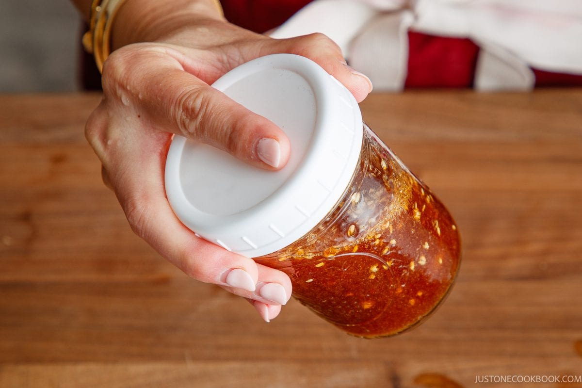 A hand holds a glass jar filled with a chunky brown wafu dressing, sealed with a white plastic lid, over a wooden surface.