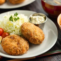 A plate with two Menchi Katsu (Ground Meat Cutlet) patties, shredded cabbage, cherry tomatoes, a lemon wedge, and a small cup of sauce. Bowls of miso soup and white rice are in the background with chopsticks beside the plate.