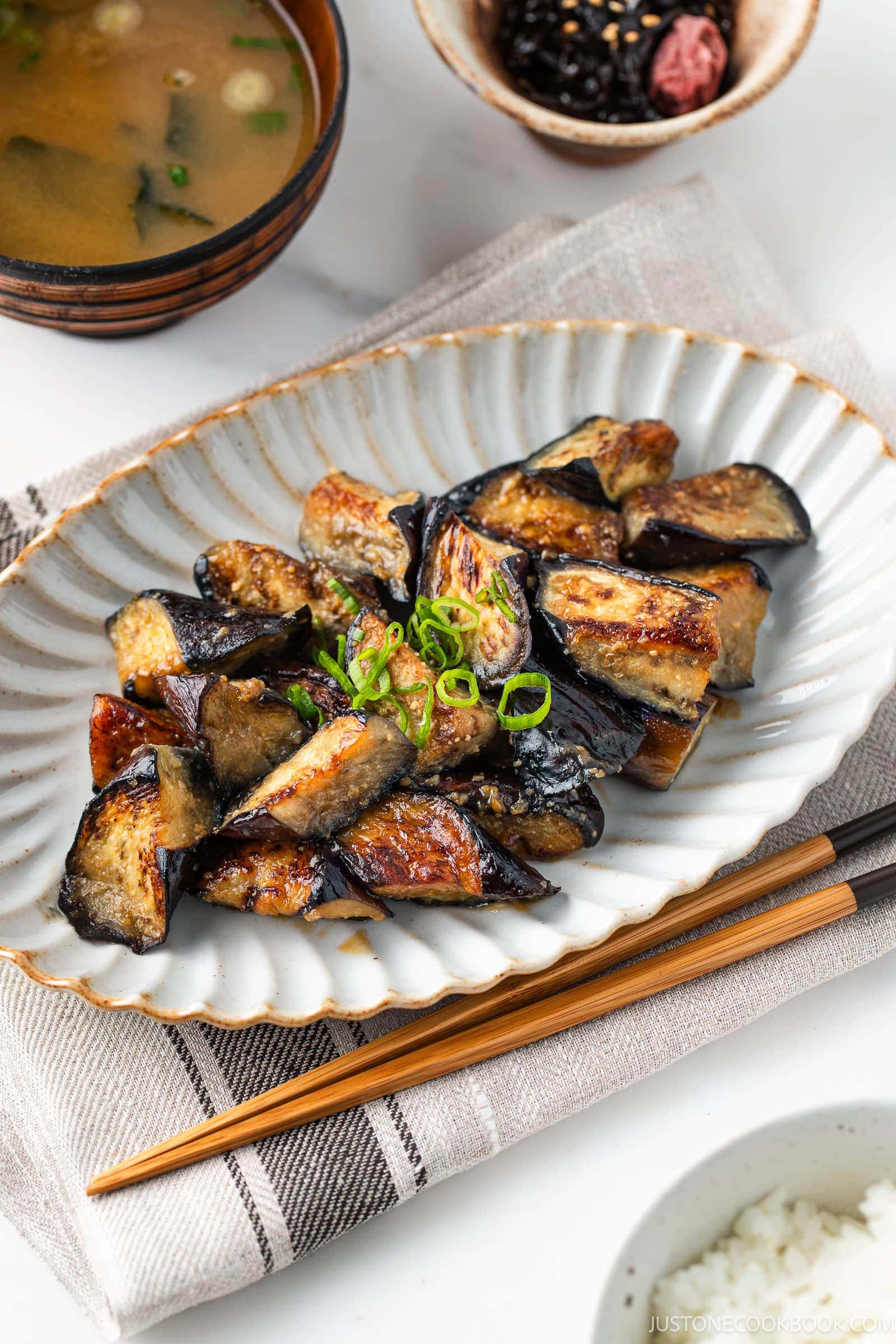 Stir-fried eggplant slices garnished with chopped green onions on a ceramic plate evoke the savory flavors of a Miso Eggplant Stir-Fry, accompanied by chopsticks, a bowl of miso soup, and a small dish of pickles in the background.