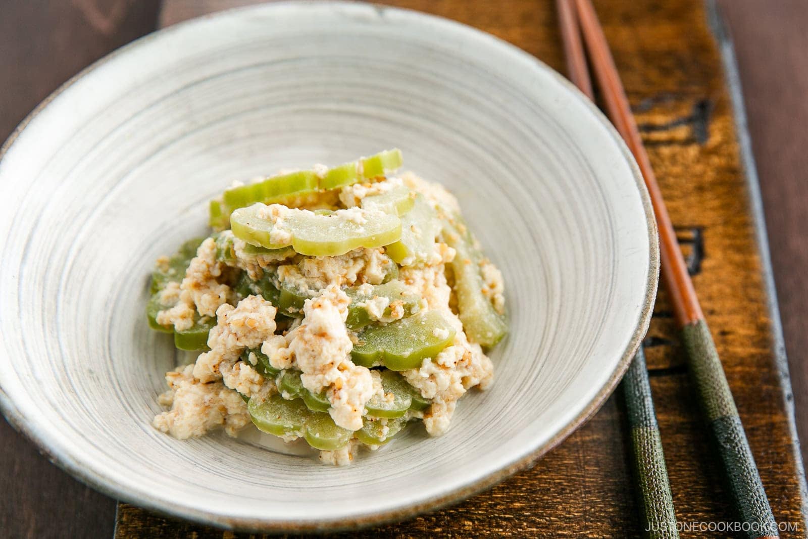 A white bowl contains a dish of sliced green bitter melon mixed with mashed tofu, reminiscent of the freshness found in a Bitter Melon and Tofu Salad. Chopsticks rest on the side of the bowl, which sits on a wooden surface.