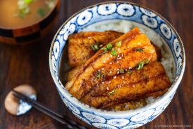 A bowl of steamed rice topped with glazed catfish kabayaki fillets, garnished with sesame seeds and chopped green onions, served with a bowl of soup and chopsticks on the side.