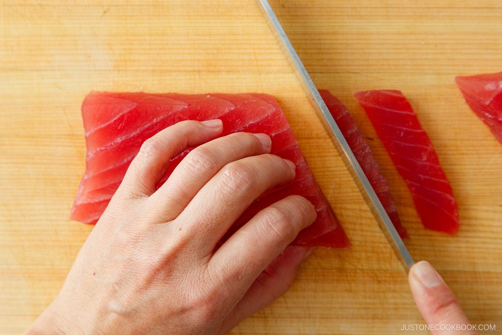 A hand demonstrates how to cut sashimi by holding a block of raw tuna and slicing it with a knife on a wooden cutting board.