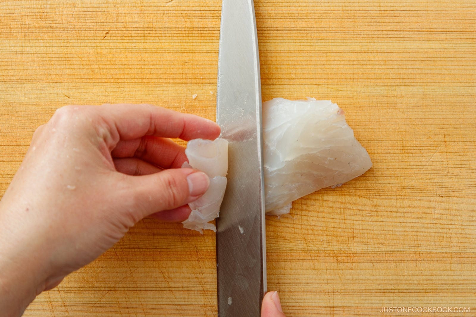 A hand demonstrates how to cut sashimi, expertly slicing a raw white fish fillet with a large knife on a wooden cutting board.