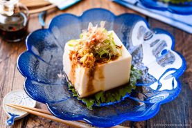 A block of chilled Hiyayakko (Japanese Cold Tofu) topped with green onions, grated ginger, bonito flakes, and soy sauce sits on a shiso leaf in a blue glass dish, with chopsticks and sauce nearby.