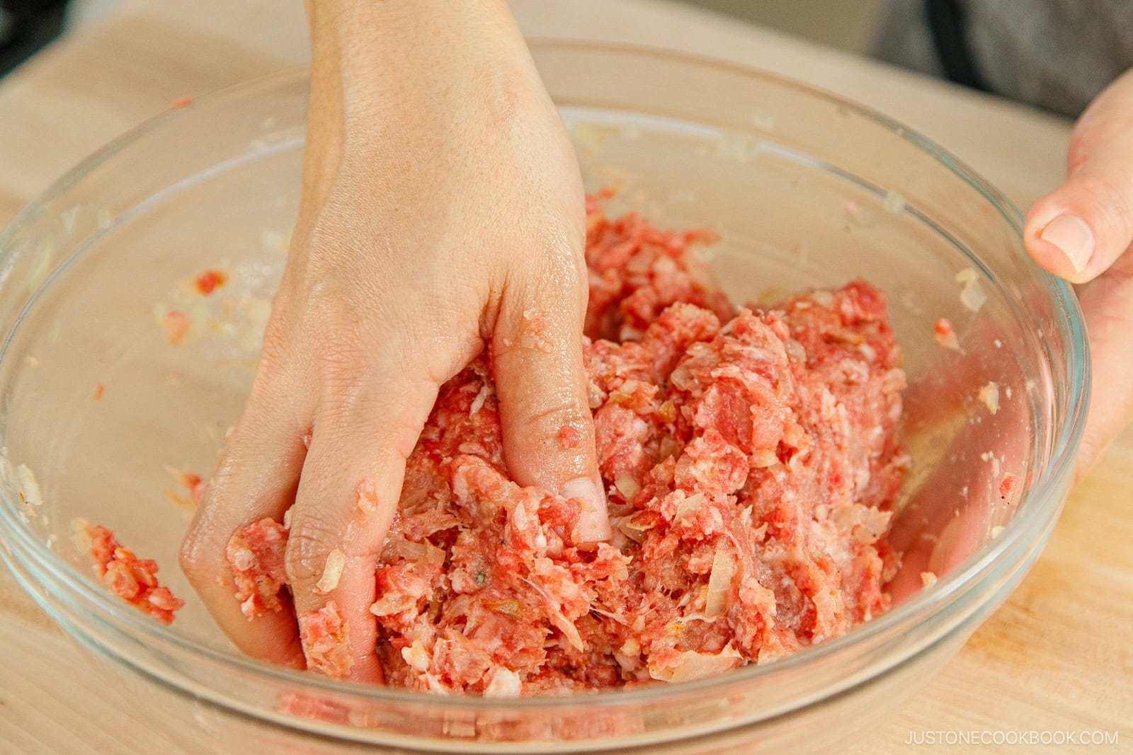 A hand mixes raw ground meat with chopped onions and seasonings in a clear glass bowl on a wooden surface, preparing the filling for menchi katsu.
