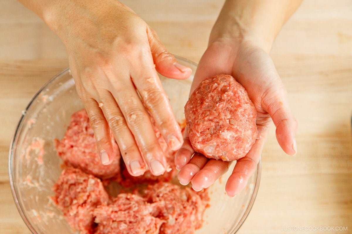Hands shaping a portion of raw ground meat mixture into a patty for menchi katsu over a bowl filled with more of the same meat mixture, on a light wooden surface.