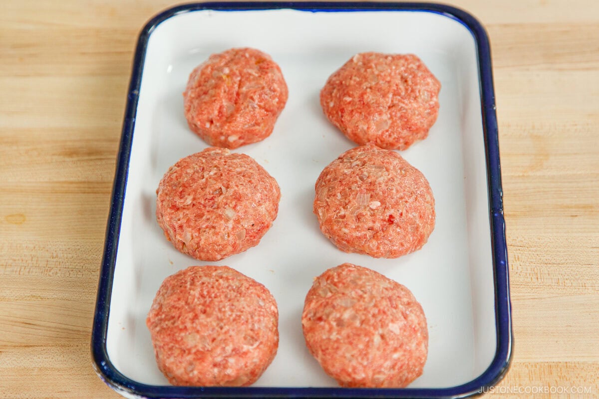 Six uncooked hamburger patties, perfect for making menchi katsu, are arranged in two rows on a white rectangular tray with a blue rim, placed on a wooden surface.