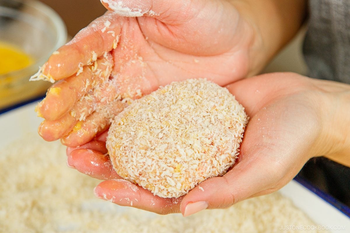 A close-up of hands holding a menchi katsu patty, being coated in panko breadcrumbs, with a bowl containing more breadcrumbs in the background.