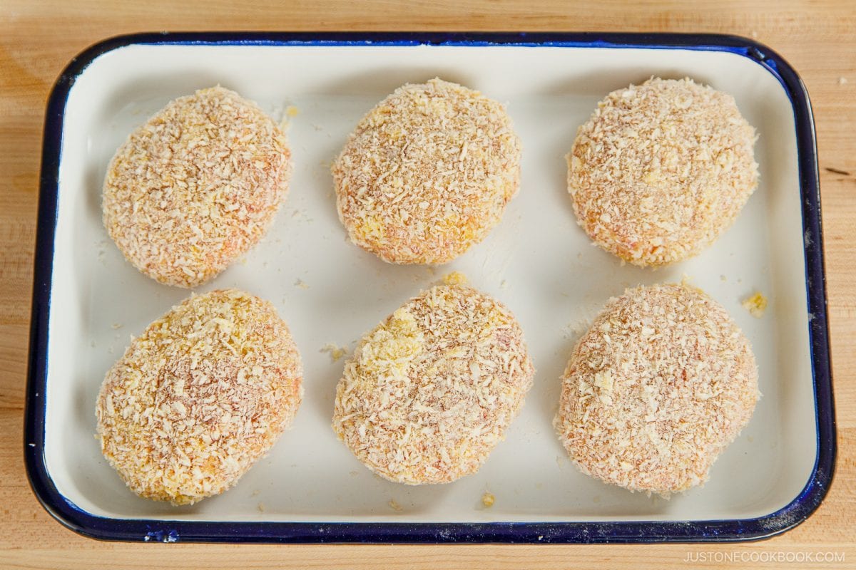 Six breaded menchi katsu patties are arranged in two rows on a rectangular white tray with a dark rim, placed on a wooden surface. The patties appear to be uncooked and coated with panko breadcrumbs.