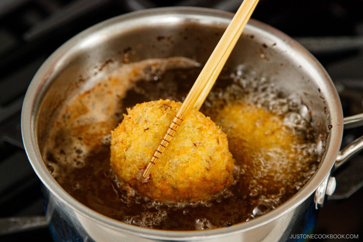A golden-brown menchi katsu is being lifted from hot oil in a saucepan with chopsticks, showing deep frying in progress.