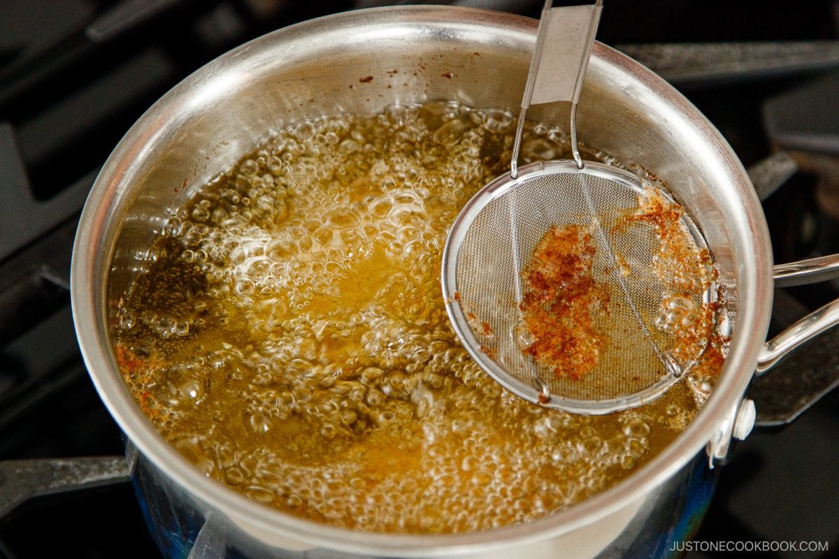 A metal strainer holds browned crumbs above a pot of hot oil, which is actively bubbling on the stovetop as menchi katsu is deep fried to crispy perfection.