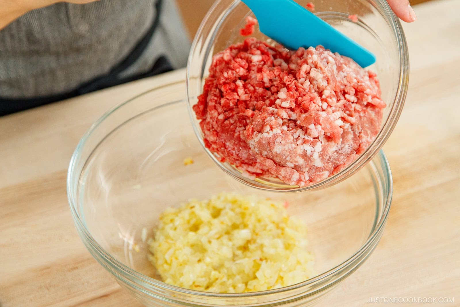 A person pours ground meat from a glass bowl into a larger bowl of chopped onions with a blue spatula, prepping the mixture for homemade menchi katsu on a wooden surface.