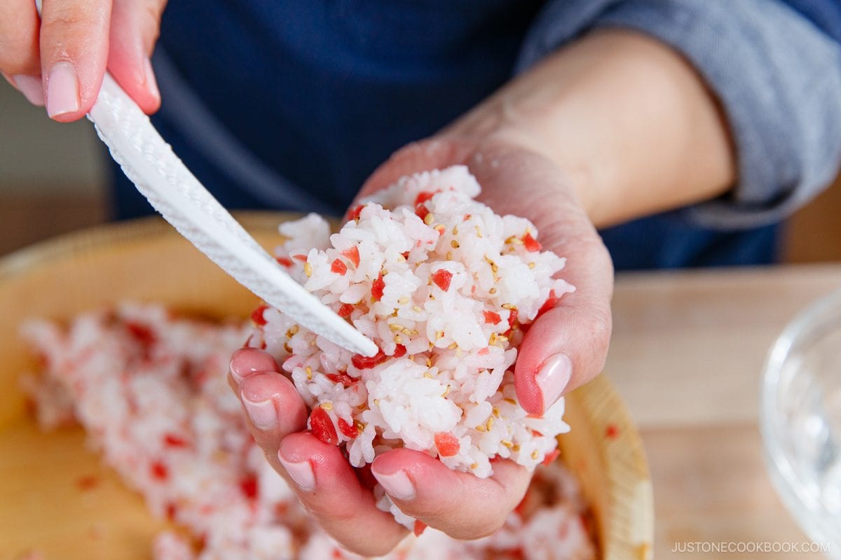 A person shapes Umeboshi Onigiri (Plum Rice Ball), mixing rice with red bits and sesame seeds in their hand, using a knife to form or cut the mixture, with a wooden bowl of rice in the background—a nod to the comforting scenes from Midnight Diner.