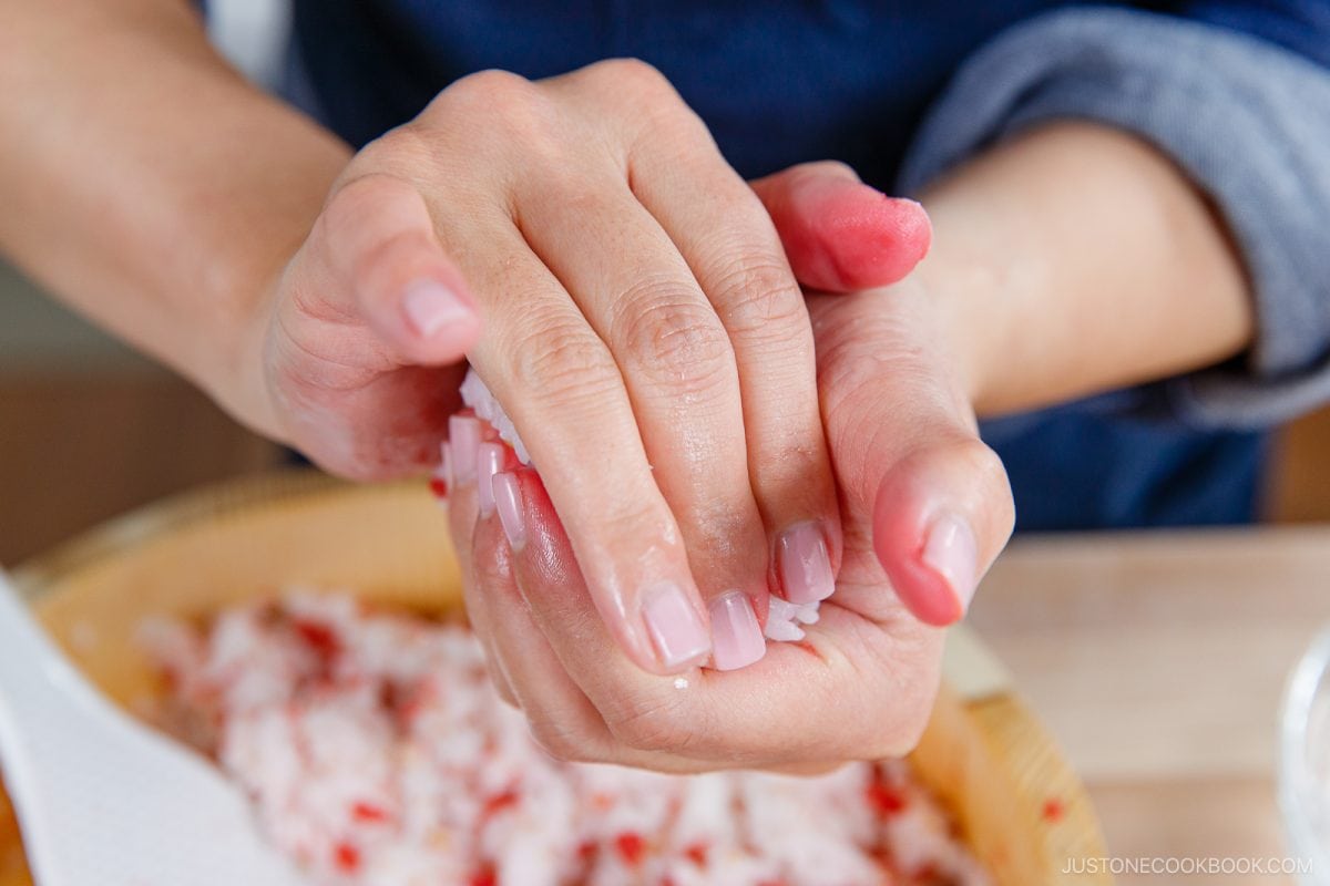 Close-up of hands shaping rice into a ball, with a bowl of rice and a spatula in the background. The person’s fingers are slightly pink, and the scene suggests making Umeboshi Onigiri (Plum Rice Ball), reminiscent of the comforting recipes featured in Midnight Diner.