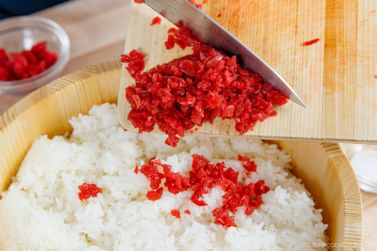 Chopped red pickles are being added from a wooden cutting board onto a bowl of white rice, reminiscent of the classic Umeboshi Onigiri (Plum Rice Ball) from Midnight Diner, with a small glass bowl visible in the background.