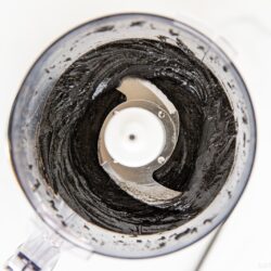 A food processor bowl filled with thick, smooth black paste, viewed from above. The sharp metal blades in the center are surrounded by the dark mixture—just like when learning how to make black sesame paste (Kuro Neri Goma).