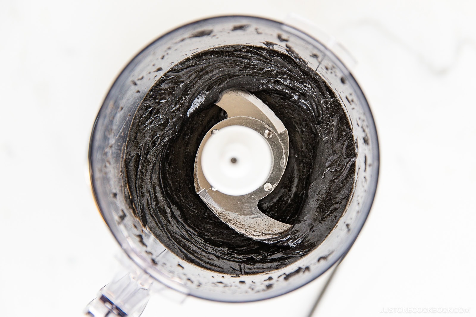 A food processor bowl filled with thick, smooth black paste, viewed from above. The sharp metal blades in the center are surrounded by the dark mixture—just like when learning how to make black sesame paste (Kuro Neri Goma).