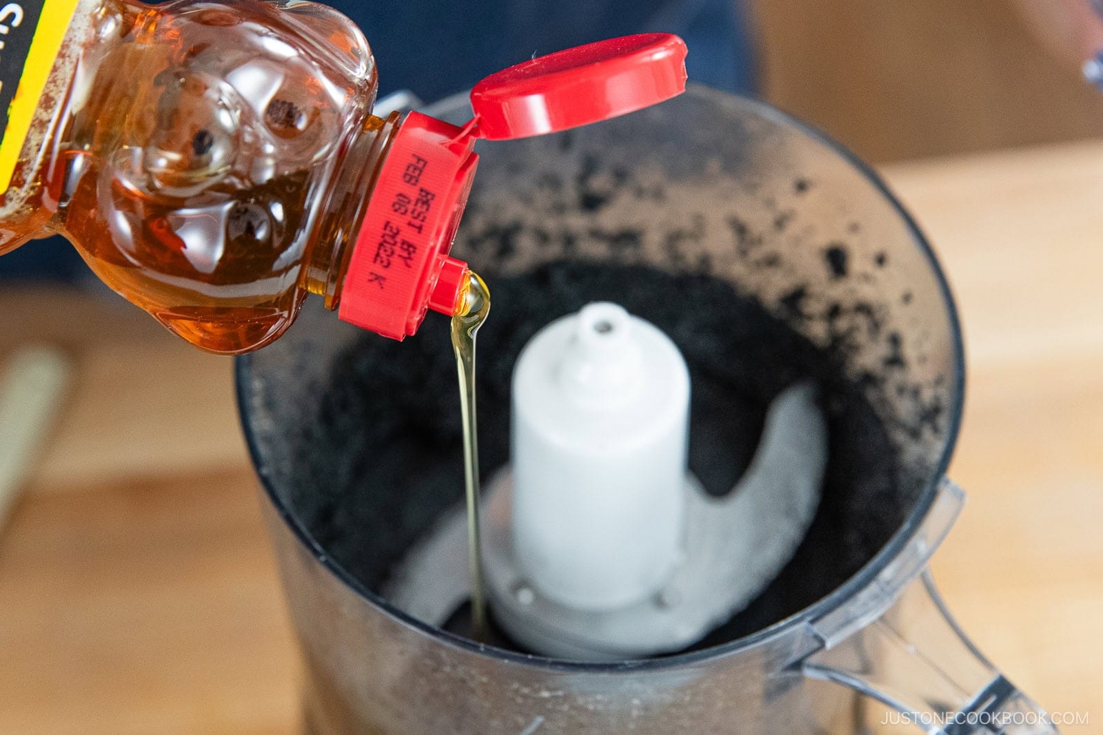 Honey is being poured from a squeeze bottle into a food processor containing ground black ingredients, possibly for How to Make Black Sesame Paste (Kuro Neri Goma) using crushed cookies or chocolate crumbs.