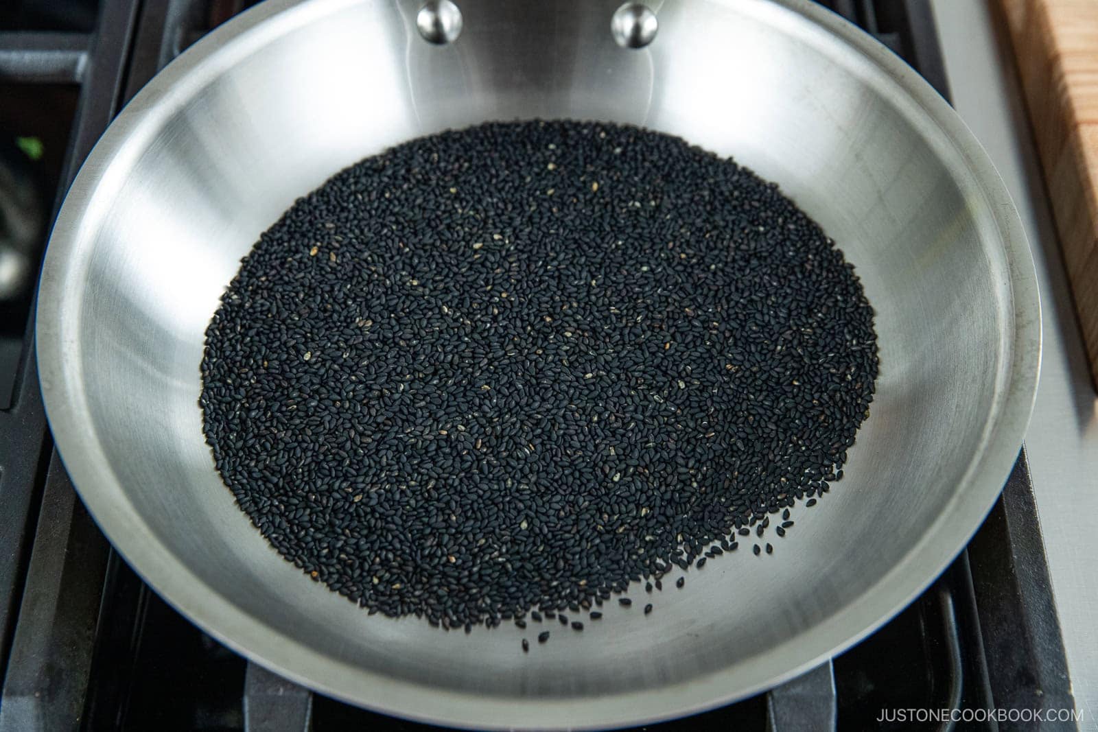 A stainless steel pan sits on a stove, filled with a layer of black sesame seeds being toasted—an essential step in How to Make Black Sesame Paste (Kuro Neri Goma). Some seeds are scattered near the edge of the pan.