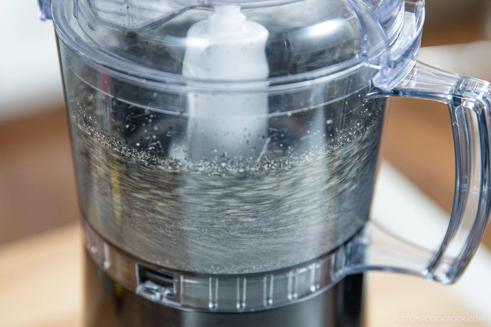 A close-up of a food processor blending water, with small bubbles and swirls forming inside the transparent container—a key step in How to Make Black Sesame Paste (Kuro Neri Goma).