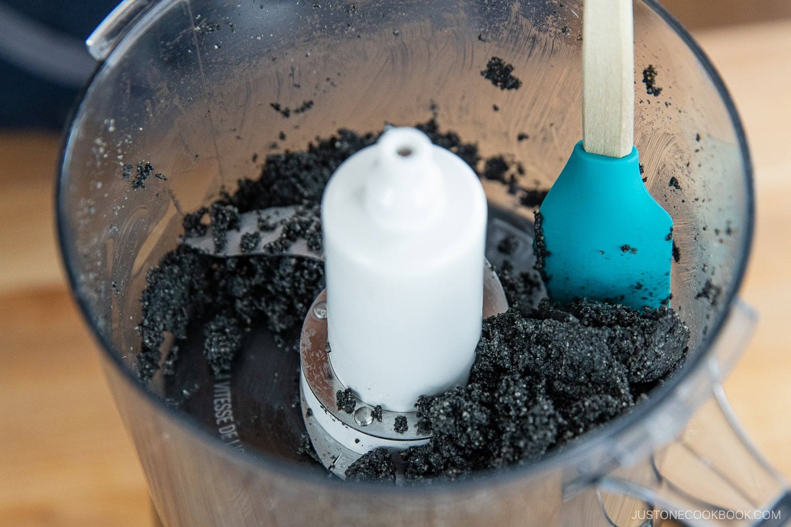 A food processor containing a coarse black paste for How to Make Black Sesame Paste (Kuro Neri Goma) with a teal spatula resting inside, seen on a wooden surface.
