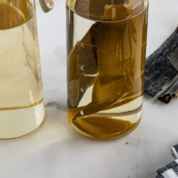 Two glass jars filled with a light yellow Kombu Dashi, one containing pieces of dried kelp. Strips of dried kelp and a checkered cloth napkin lie on a white marble surface nearby.