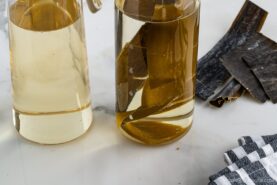 Two glass jars filled with a light yellow Kombu Dashi, one containing pieces of dried kelp. Strips of dried kelp and a checkered cloth napkin lie on a white marble surface nearby.