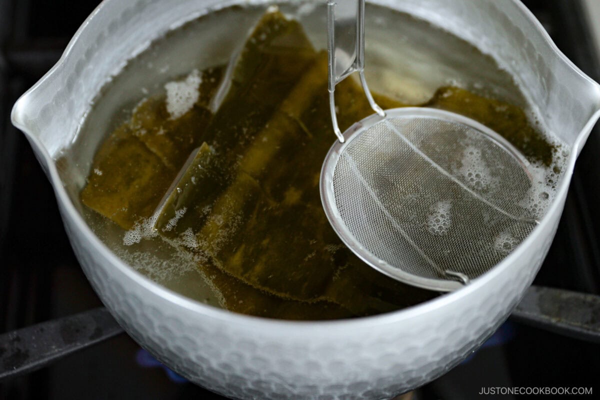 A pot of water with large pieces of kombu is being simmered to make kombu dashi, with a fine mesh strainer partially submerged, likely preparing this umami-rich broth on a stovetop.