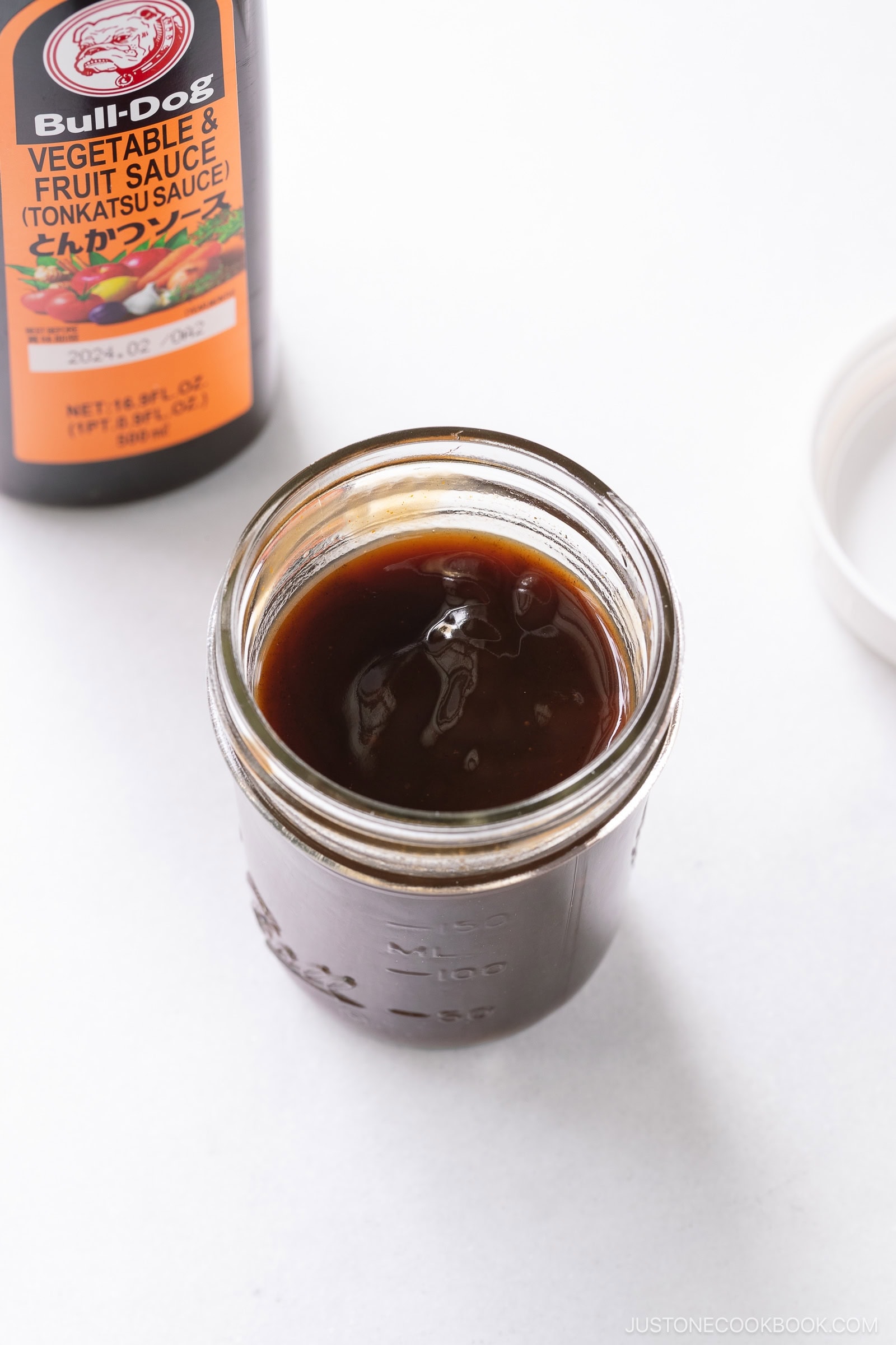 An open glass jar filled with dark brown tonkatsu sauce, made from a classic Tonkatsu Sauce Recipe, sits on a white surface next to a bottle labeled Bull-Dog Vegetable & Fruit Sauce. The jar&rsquo;s lid is off and placed nearby.