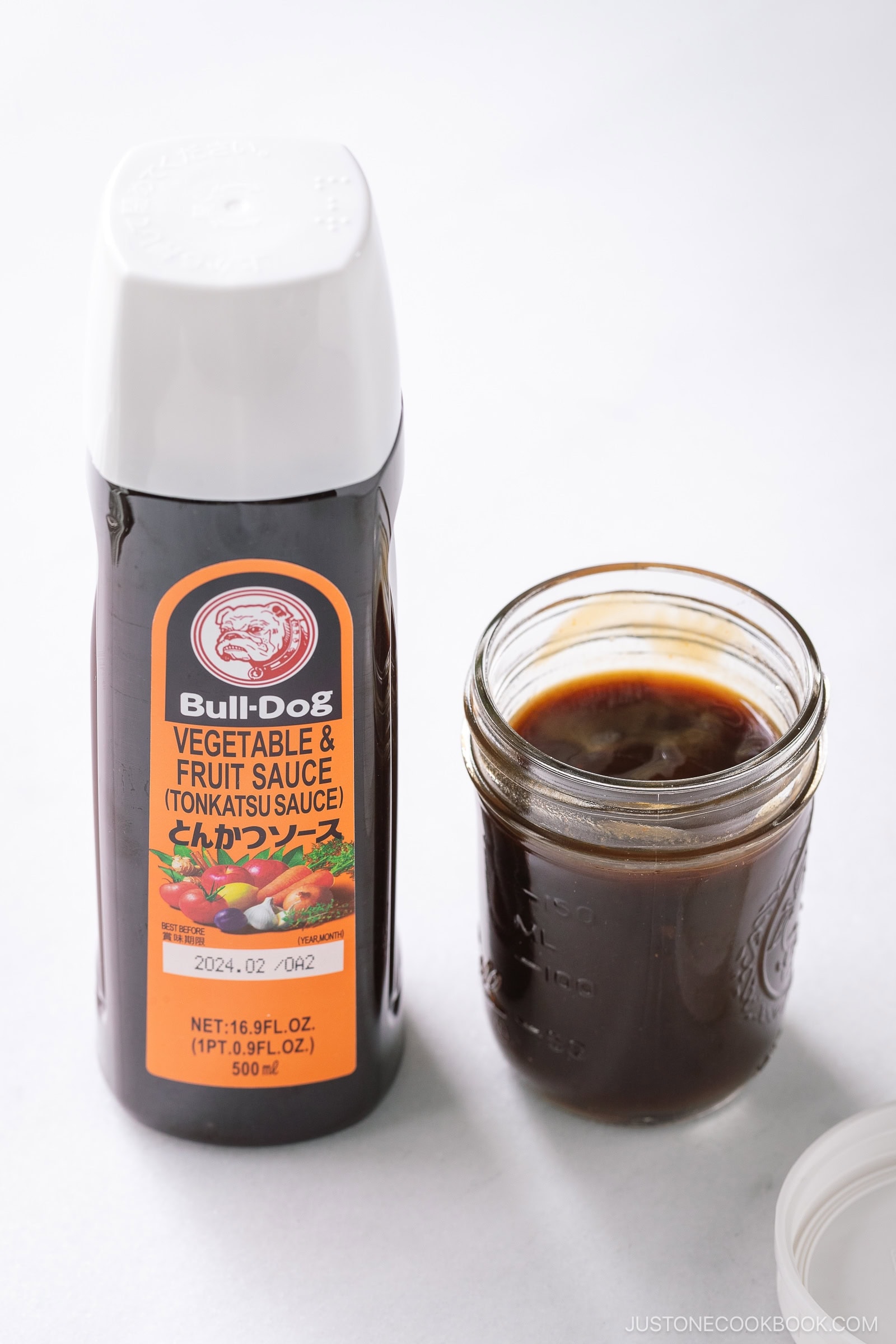 A bottle of Bull-Dog Vegetable & Fruit Sauce (Tonkatsu sauce), perfect for any Tonkatsu Sauce recipe, stands next to an open glass jar filled with dark brown sauce on a white surface.