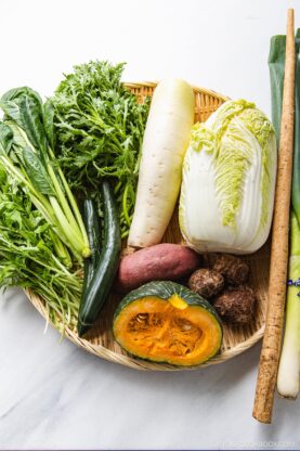 A bamboo basket filled with unique Japanese vegetables, including napa cabbage, daikon radish, kabocha squash, burdock root, sweet potato, cucumber, taro, leek, and leafy greens on a white surface.