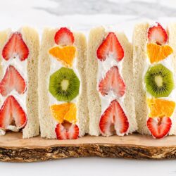 Four Japanese Fruit Sandwiches with cream, filled with slices of strawberries, oranges, and kiwis, are neatly arranged on a wooden board against a light background.