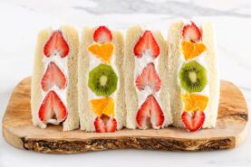 Four Japanese Fruit Sandwiches with cream, filled with slices of strawberries, oranges, and kiwis, are neatly arranged on a wooden board against a light background.