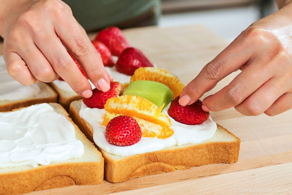 Close-up of hands assembling a Japanese Fruit Sandwich, placing strawberries, orange slices, and kiwi on cream-covered white bread, with another slice beside it on a wooden surface.