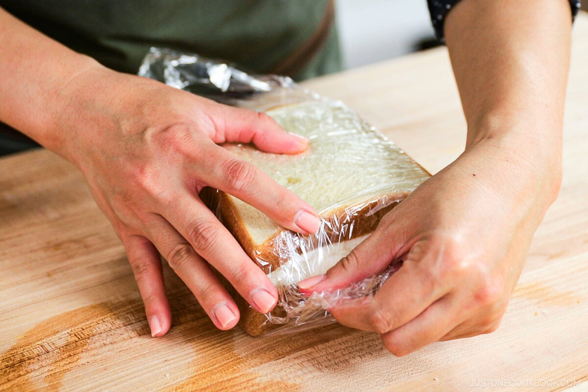 A person wraps a slice of white bread in plastic wrap on a wooden surface, using both hands to secure the wrap&mdash;preparing the base for a delicious Japanese Fruit Sandwich.