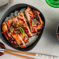 A bowl of white rice topped with sliced miso chicken, garnished with sesame seeds and sliced green onions. Chopsticks and a green glass are nearby on a light-colored tablecloth.