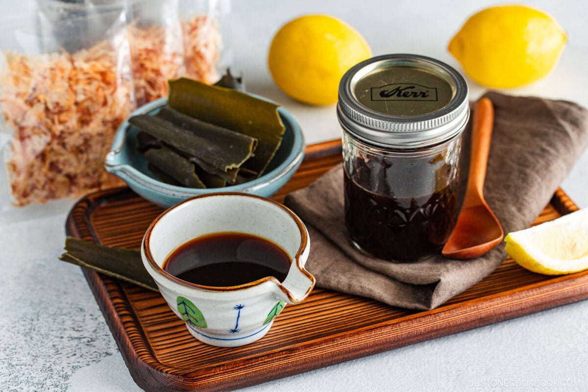 A tray with a jar of dark ponzu sauce, a small bowl showcasing the Ponzu Sauce Recipe, dried seaweed strips, dried flakes, a wooden spoon, a lemon wedge, and two whole lemons in the background.