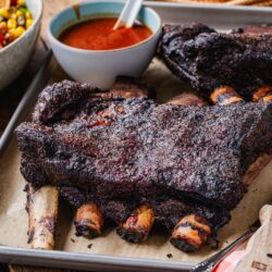 A tray with large, smoked beef ribs on parchment paper, a bowl of barbecue sauce with a spoon, a knife and fork, and a bowl of colorful corn salad sit on a rustic wooden table. The smoked beef ribs are the star of this hearty spread.
