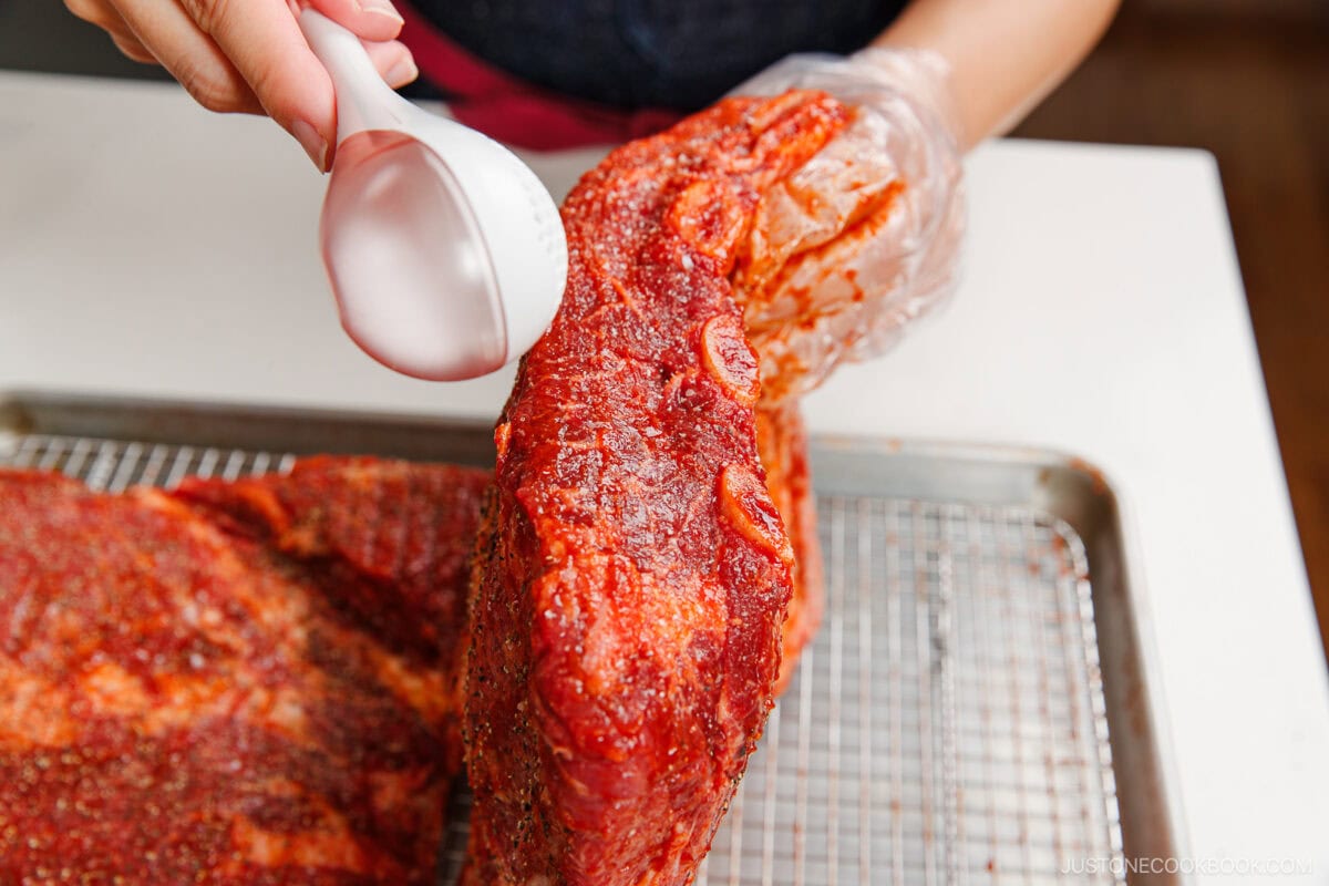 A person wearing a glove sprinkles liquid over a large, seasoned piece of raw smoked beef ribs placed on a wire rack above a tray.