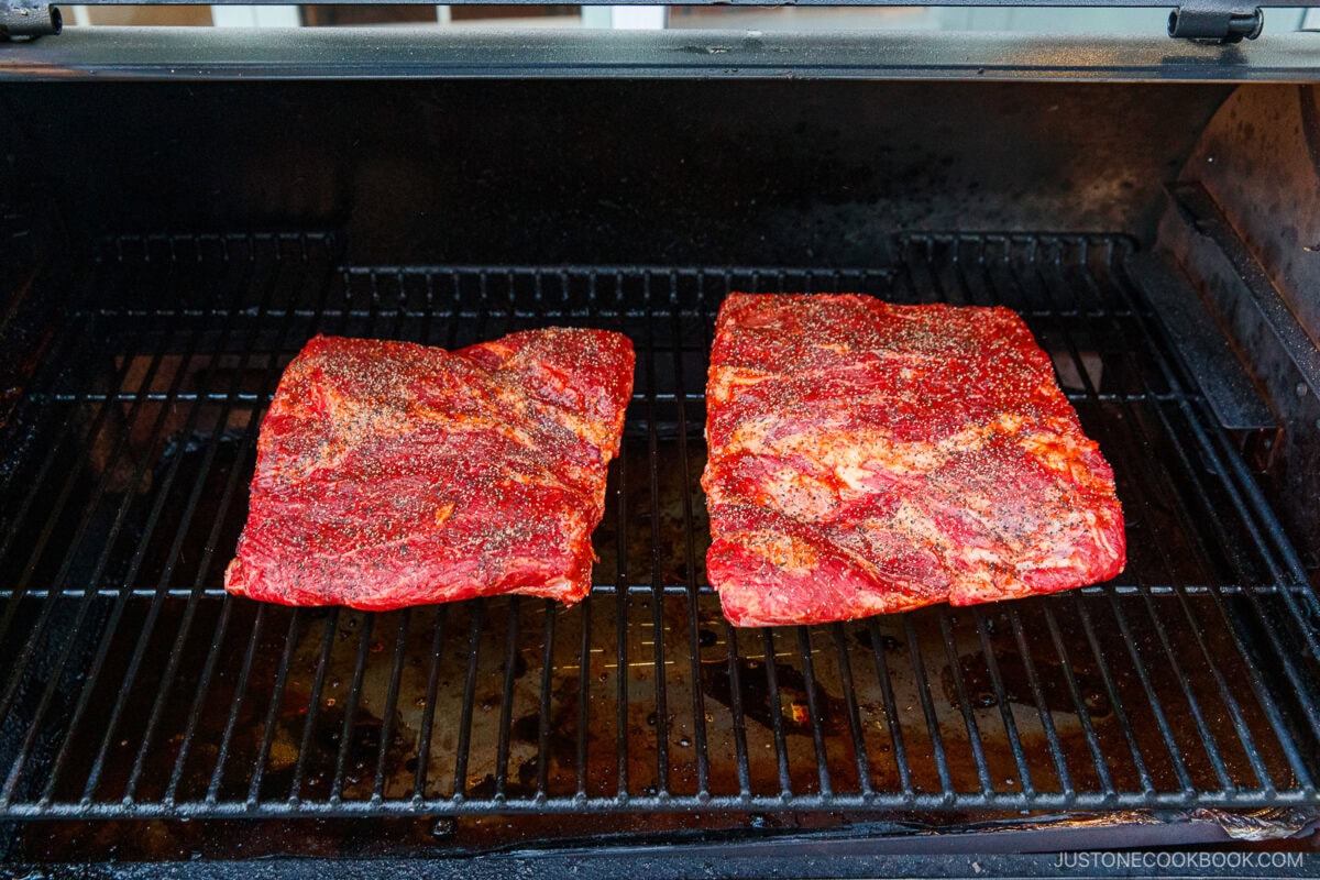Two large, seasoned brisket cuts and smoked beef ribs rest on the grates of an open barbecue smoker, soaking up rich, smoky flavor.