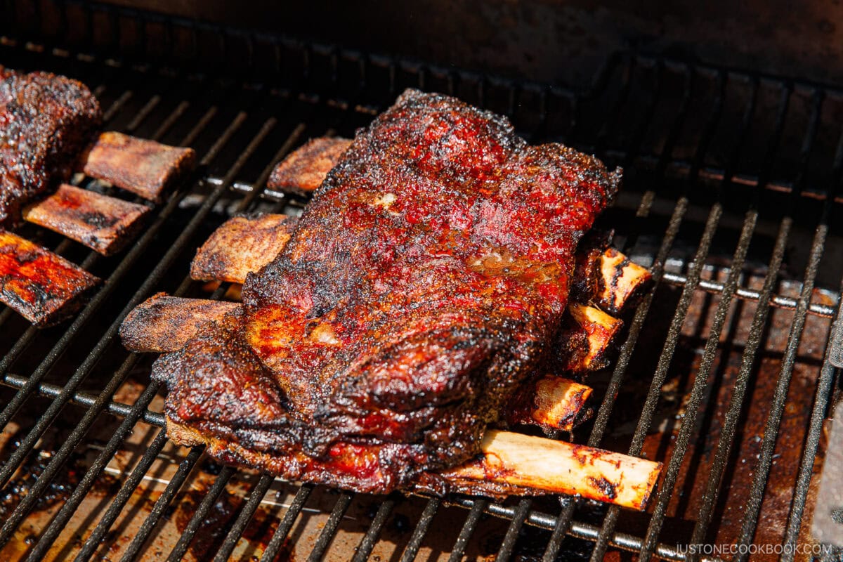 Smoked beef ribs with a dark, crispy, and seasoned crust are cooking on a grill, their char marks and juicy texture glistening under bright sunlight.