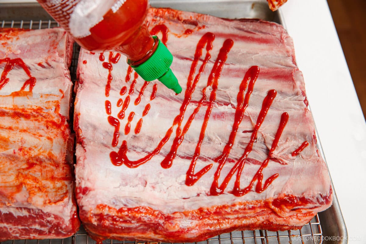 A hand squeezes sriracha sauce onto large, raw pork ribs placed on a wire rack over a baking sheet, evoking the mouthwatering preparation often seen with smoked beef ribs.
