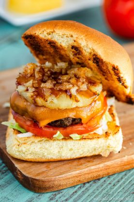 The Teriyaki Burger features a beef patty, cheddar cheese, grilled pineapple, chopped onions, tomato slices, lettuce, and sauce on a toasted bun, all served on a wooden board.