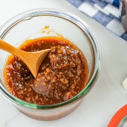 A glass jar filled with chunky brown All Purpose Miso Sauce, stirred with a wooden spoon, sits on a white surface next to a blue-striped cloth and a red-lidded jar.