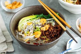 A bowl of Black Sesame Dan Dan Noodles topped with minced meat, bok choy, bean sprouts, sliced green onions, garlic chips, and sesame seeds, with chopsticks resting on the bowl.