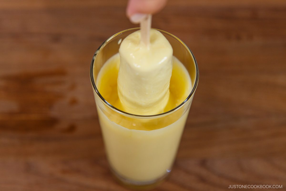 A hand dips a creamy yellow popsicle into a glass of vibrant juice, set on a wooden surface&mdash;reminiscent of the playful, inventive spirit behind treats like the Japanese Corn Dog.