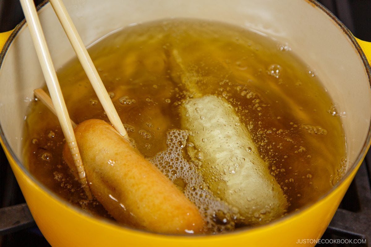 Two Japanese Corn Dogs are being deep-fried in hot oil inside a yellow pot, with one corn dog being turned using wooden chopsticks. The oil is bubbling around the corn dogs.