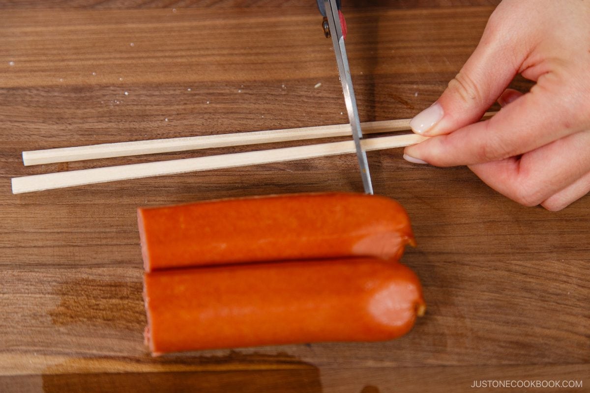 A hand uses scissors to cut two wooden chopsticks on a wooden surface, preparing them for assembling Japanese Corn Dog snacks. Two halved sausages lie nearby.