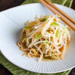A small pile of seasoned Bean Sprout Salad garnished with sliced green onions on a white plate, with chopsticks resting on the side and a green cloth underneath.