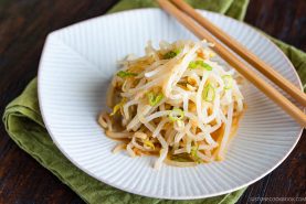 A small pile of seasoned Bean Sprout Salad garnished with sliced green onions on a white plate, with chopsticks resting on the side and a green cloth underneath.